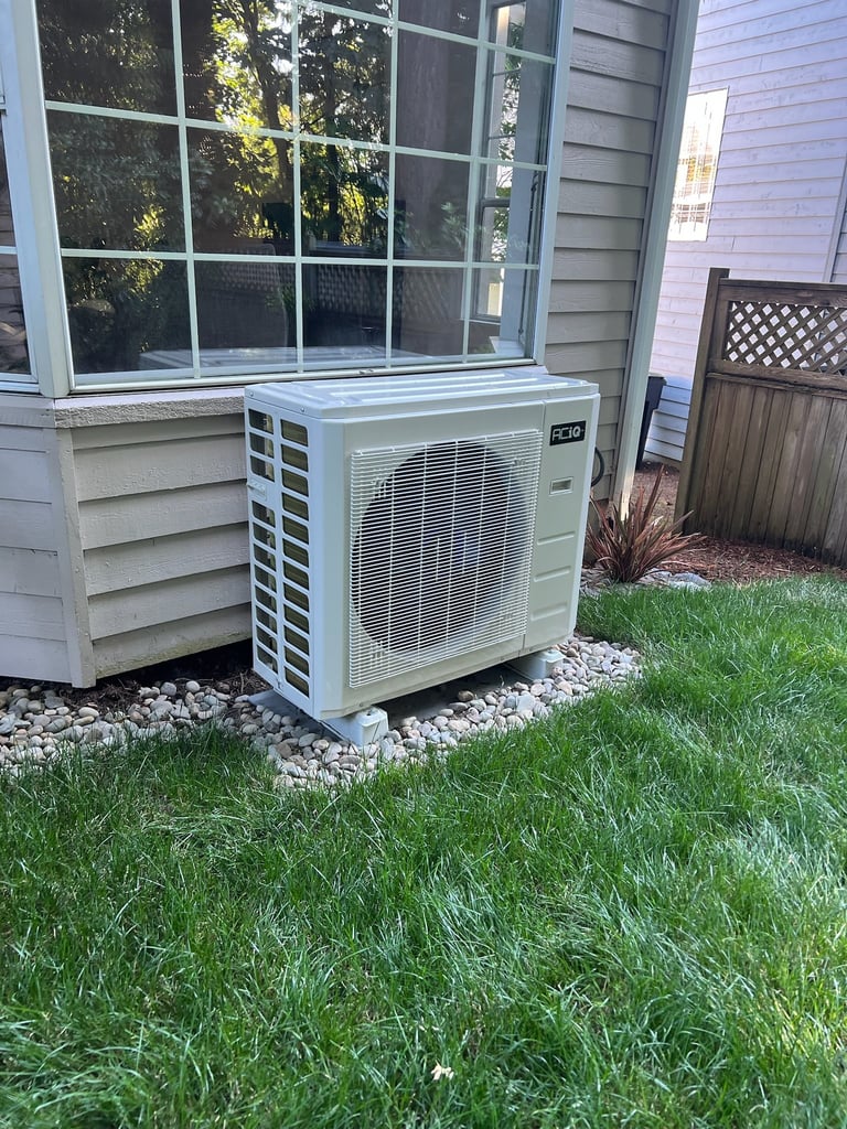 Heat pump outdoor condenser unit mounted on the exterior of a white house with a large window, surrounded by gravel and grass with fencing visible