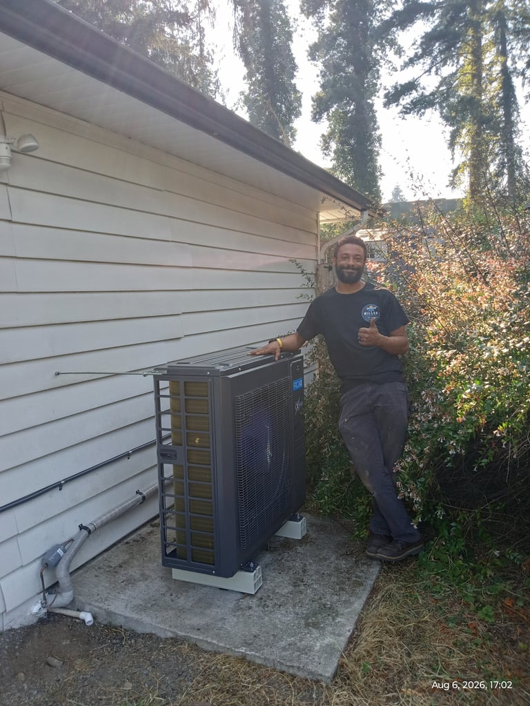 Technician standing beside a heat pump unit installed against a white house siding, with ivy-covered trees in background